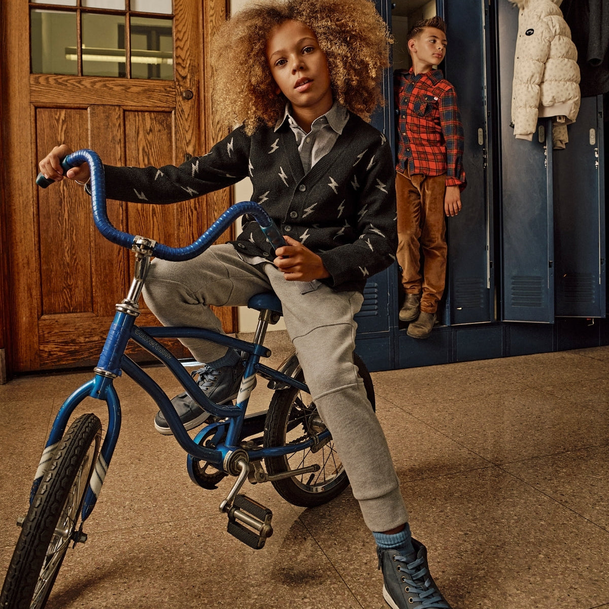 Child on a blue bicycle in a school hallway with lockers and another child in the background.