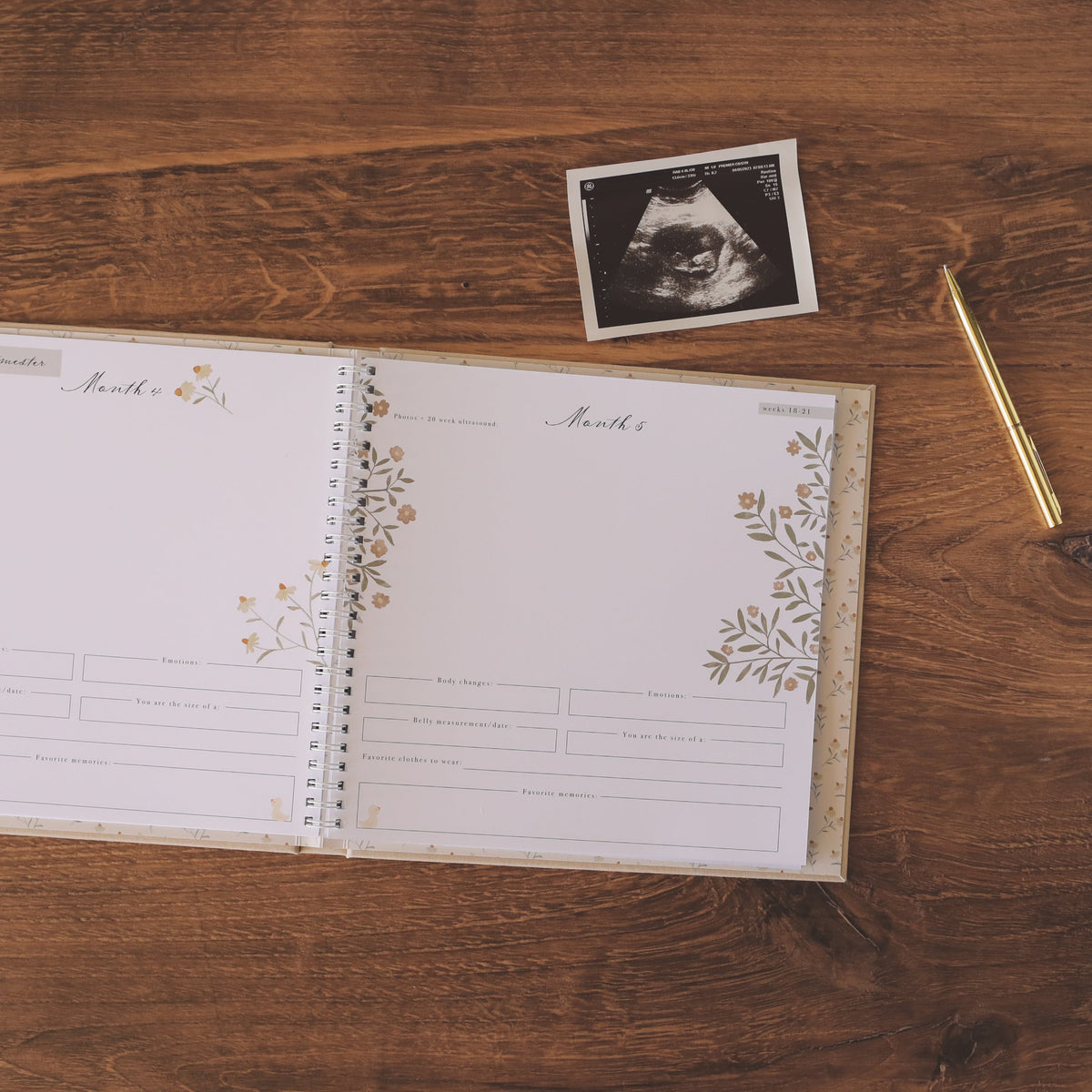 An open baby keepsake album with floral gold and beige borders lies on a wooden tabletop, alongside a small black-and-white ultrasound photo and a gold pen.
