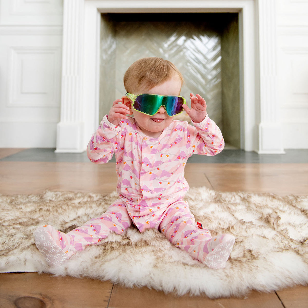 Baby in pink outfit sitting on a rug with colorful goggles