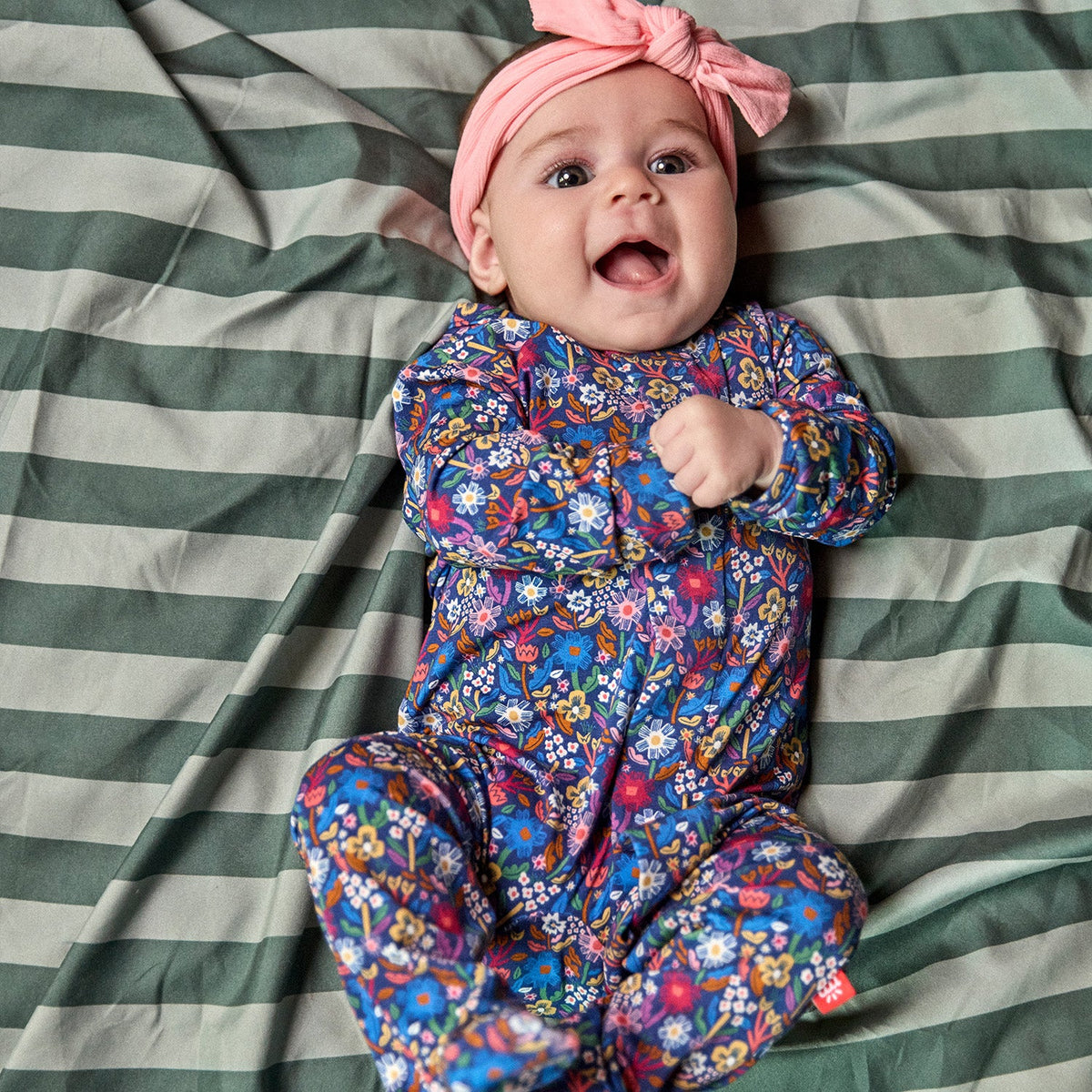 A baby lying on a green and white striped blanket, wearing a pink knotted headband and a long-sleeve footed sleeper with a colorful floral pattern.