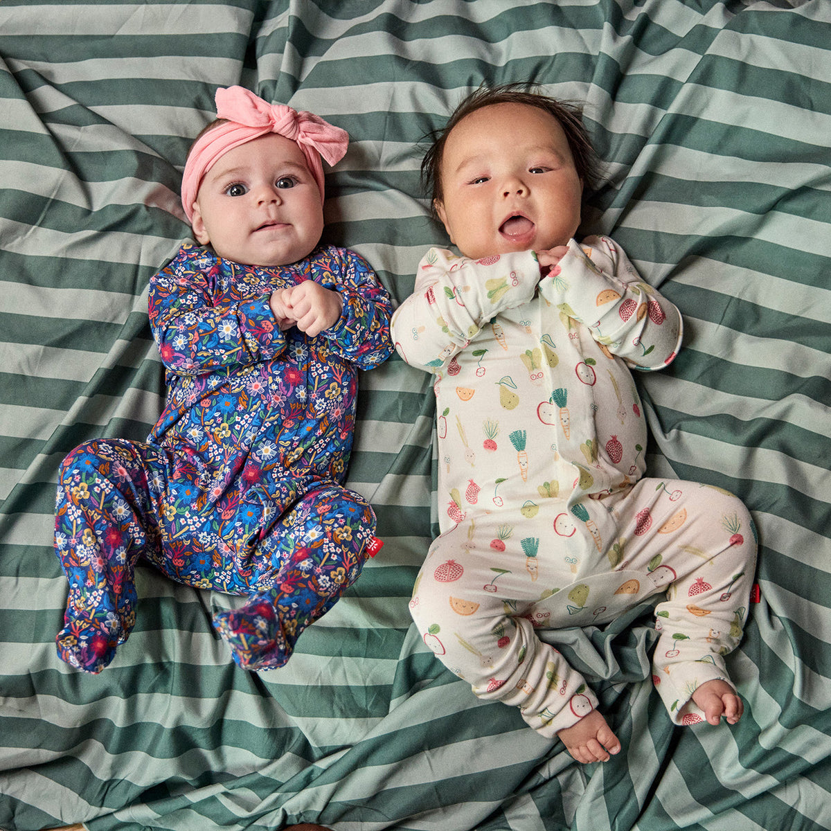 Two babies lying side by side on a green and white striped blanket, wearing matching floral and fruit-print footed onesies. The baby on the left wears a blue, purple, and pink floral one-piece with a pink bow headband.