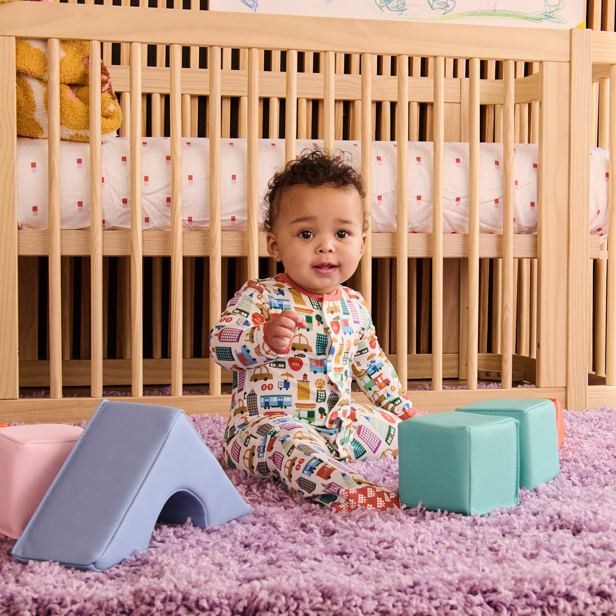A baby wearing a colorful print footed sleeper sits on a purple shag rug in front of a wooden crib, surrounded by pastel foam play blocks.