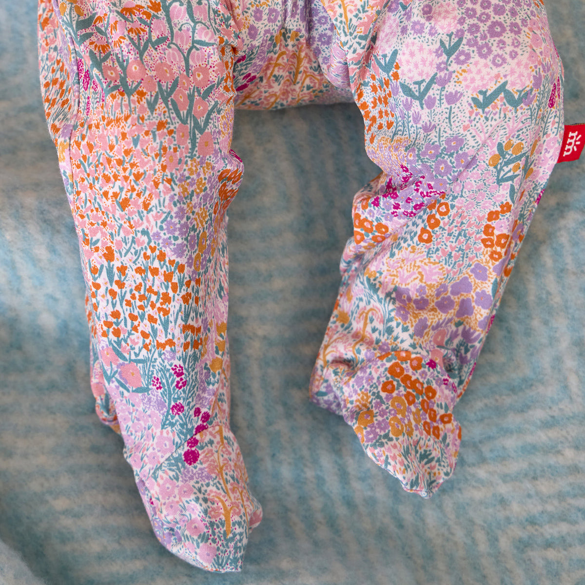 A close-up of a pair of patterned pants laid flat on a textured blue-gray surface. The fabric has an all-over floral and botanical print in pink, purple, orange, teal, and white, with a small red brand tag visible on the upper right leg.