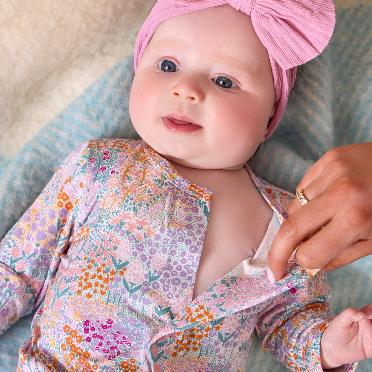 A close-up lifestyle shot of a baby wearing a pink ribbed bow headband and a pastel floral long-sleeve romper, with an adult hand adjusting the front of the romper.