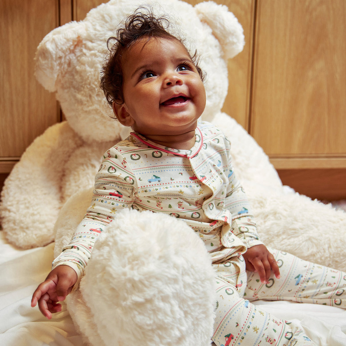 A smiling baby wearing a cream-colored patterned pajama set sits on a large white teddy bear in front of a wooden background.