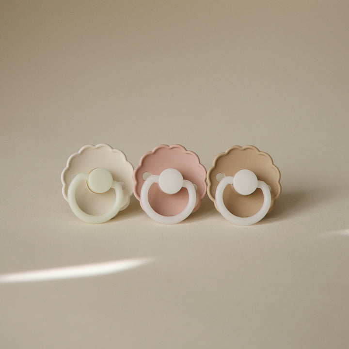 Three baby pacifiers in white, pink, and beige on a beige background