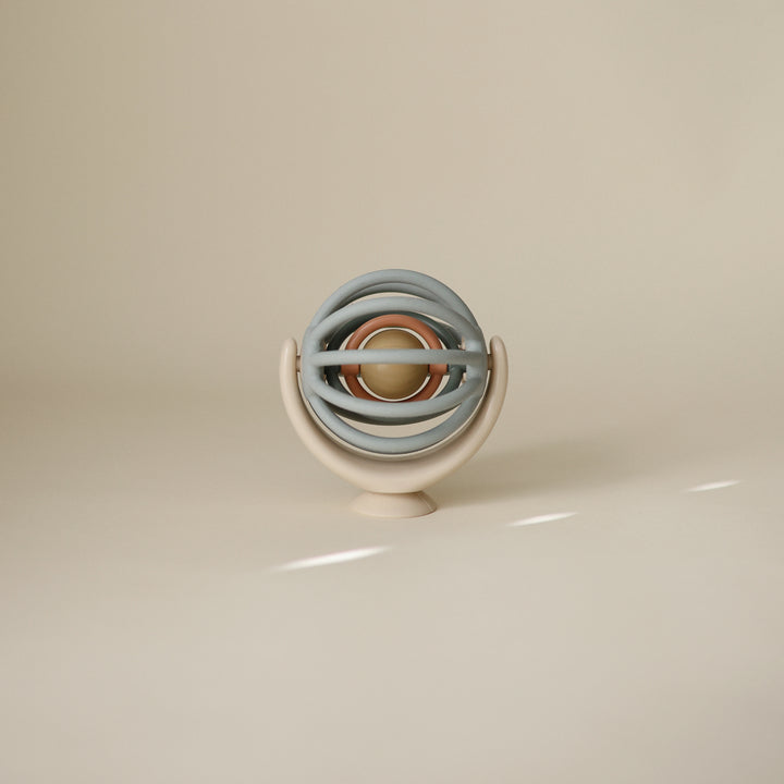A Mushie Suction Spinner Ball featuring nested pastel sage green rings and an orange center sphere, attached to a curved beige stand with a suction base against a neutral background.