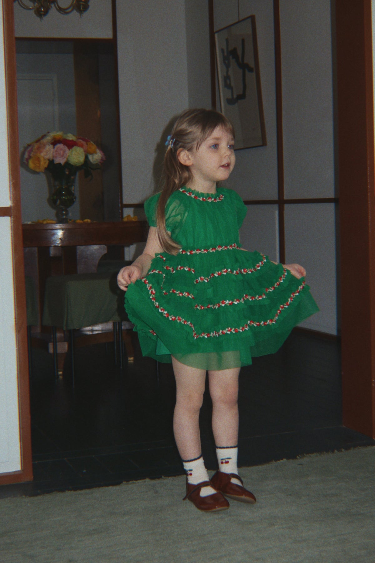 A young girl wearing a green tiered dress with colorful sequined trim, white socks, and brown shoes, standing in a doorway of an indoor room.