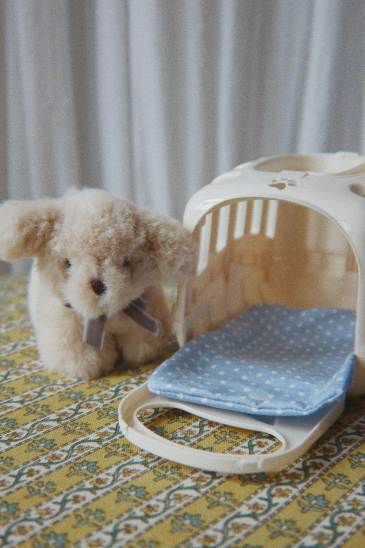 A small beige plush dog toy sits beside a cream-colored pet carrier with a blue polka-dot cushion, placed on a yellow patterned surface against a soft gray curtain background.