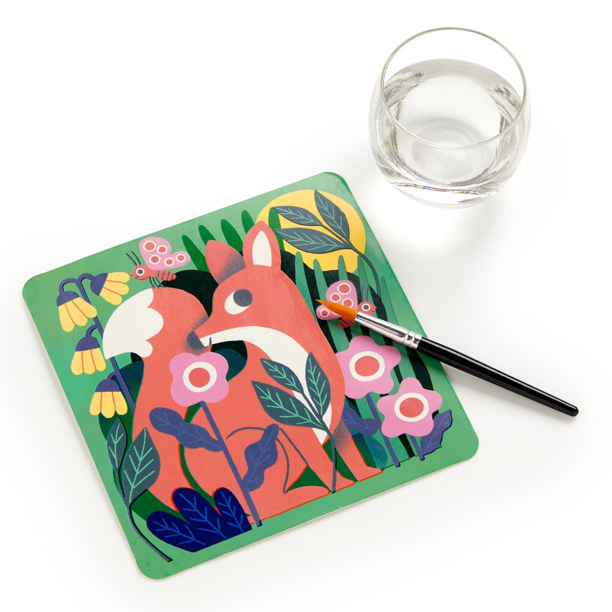 A child's water-reveal activity board featuring a colorful orange fox surrounded by pink and white flowers, green leaves, and dark branches. A paintbrush rests diagonally across the board, next to a small clear cup of water on a white background.