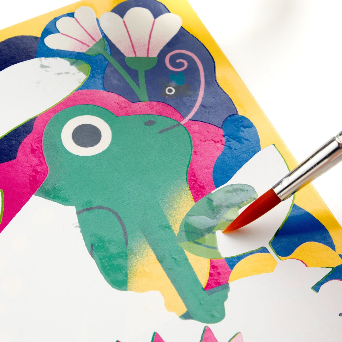 A water-reveal activity board featuring a colorful green frog surrounded by pink and white flowers, blue water details, and a dark background. A paintbrush with a red tip rests diagonally across the board, being used to paint water onto the page.