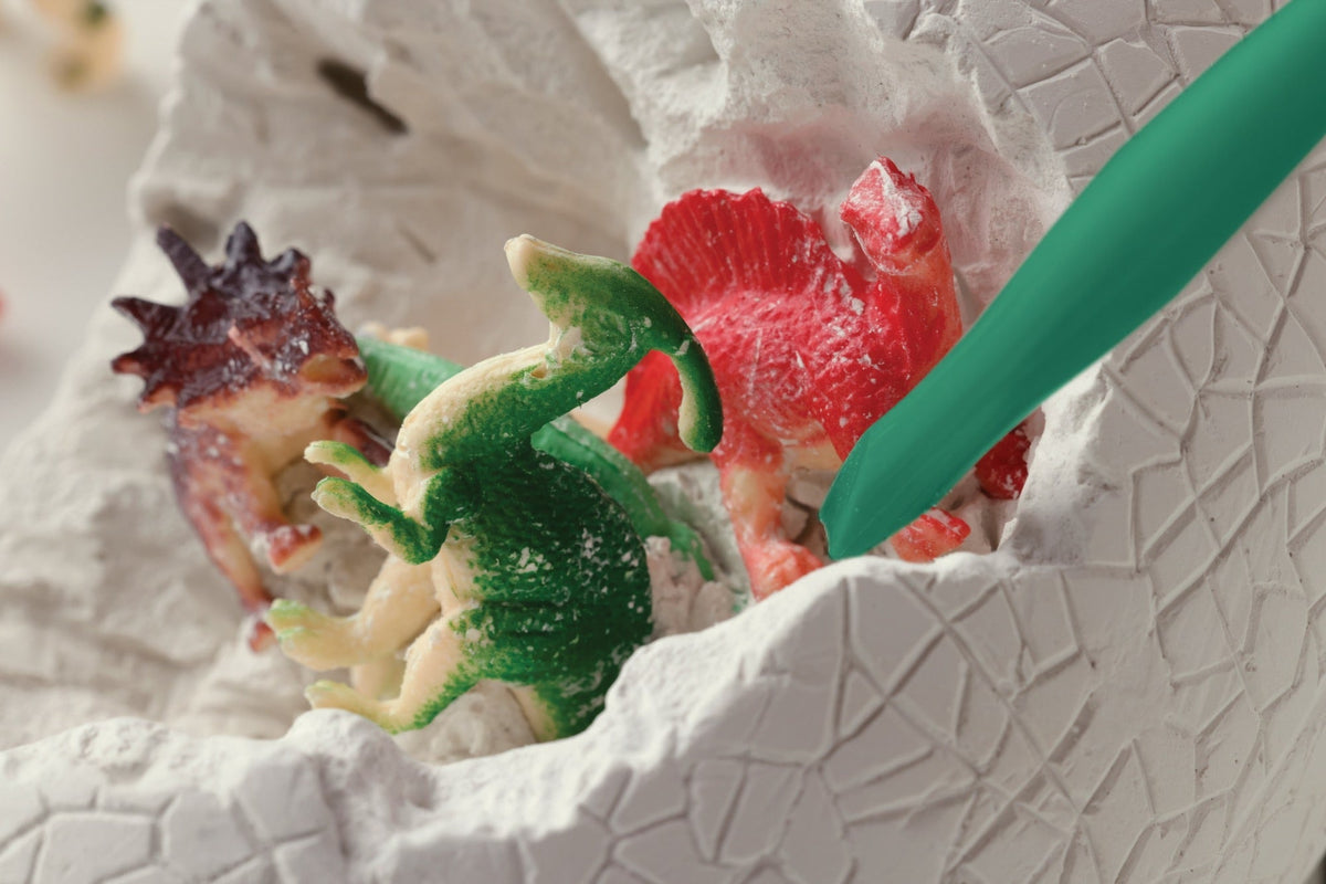 A green digging tool gently chipping away at a cracked white dinosaur egg to reveal small toy dinosaurs nestled inside.