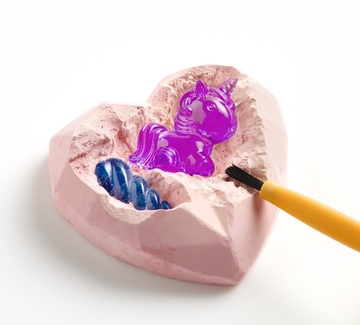 A pink heart-shaped digging block from an excavation kit, showing a partially unearthed purple unicorn figurine and blue gem pieces being brushed with a small tool.