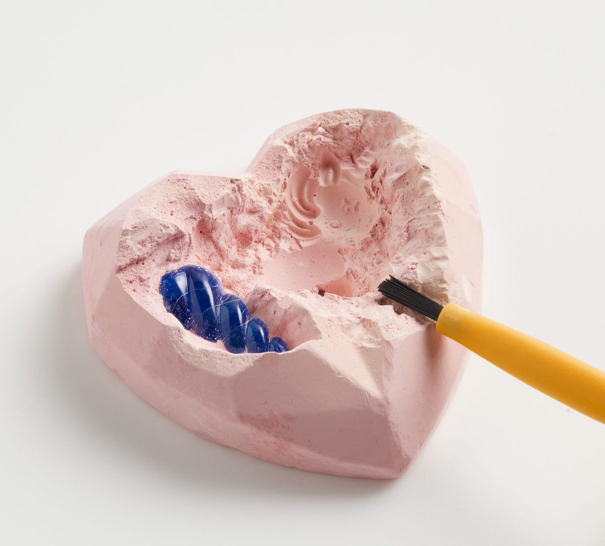 A pink heart-shaped digging block from an excavation kit, showing a partially unearthed dark blue unicorn figurine and gem pieces being brushed with a small yellow tool.