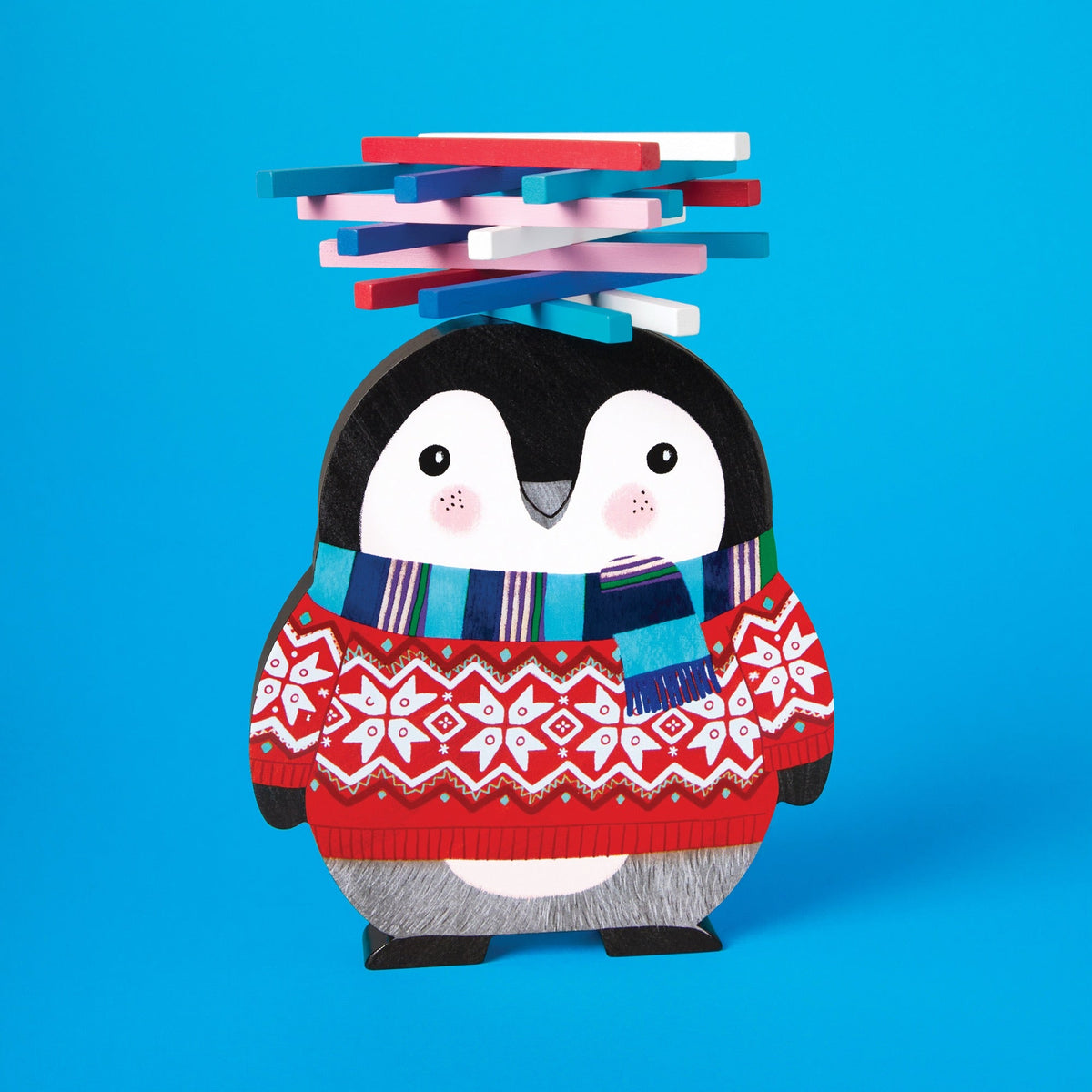 Wooden game piece for the Penguin Stack-up Wooden Balancing Game, shaped like an illustrated penguin with rosy cheeks, wearing a festive red patterned fair isle sweater and a blue-and-green striped scarf, with a balanced stack of colorful wooden sticks on its head against a bright blue background.