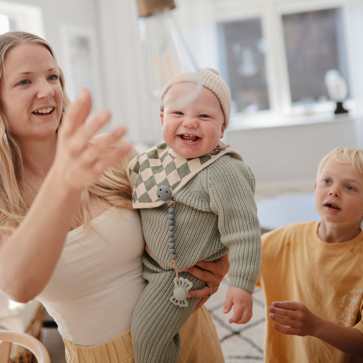 Woman holding a baby in a checkered outfit with another child in the background.