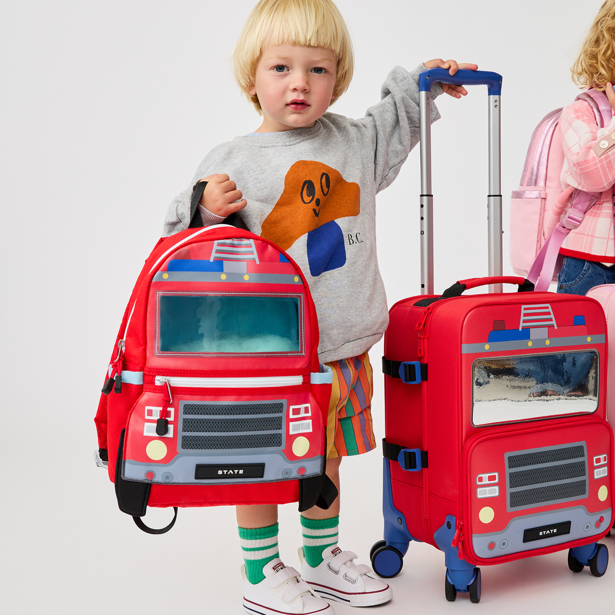 A toddler standing between a red State Bags Mini Logan firetruck-style backpack and a matching red rolling carry-on suitcase, wearing a gray sweatshirt with an orange graphic and striped socks.