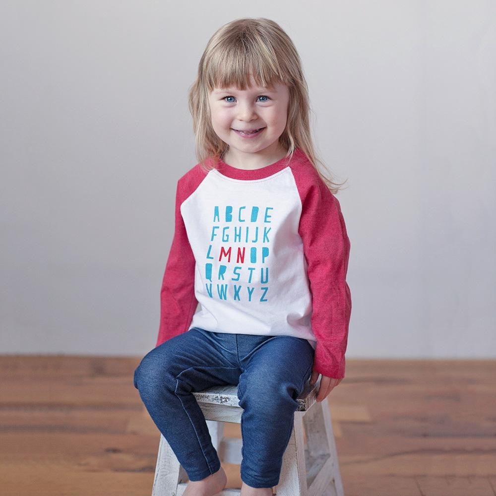 Child wearing a red and white shirt with colorful letters sitting on a stool.
