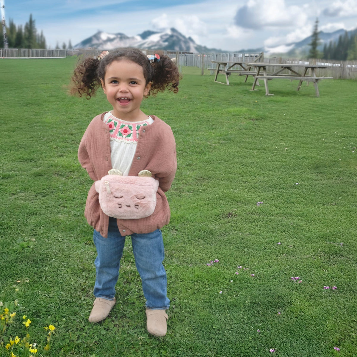 Child holding a pink bag with bear ears in a grassy field with mountains in the background