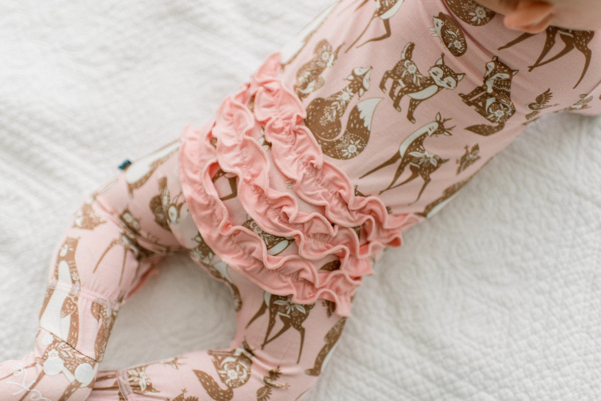 A close-up of a baby's legs wearing light pink leggings with a brown deer and fox pattern, featuring a prominent ruffled pink waistband, lying on a white textured blanket.