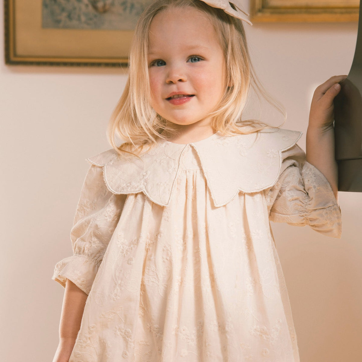 A young blonde girl wearing a cream-colored embroidered dress with a large ruffled Peter Pan collar and a matching bow in her hair, standing in an indoor setting.