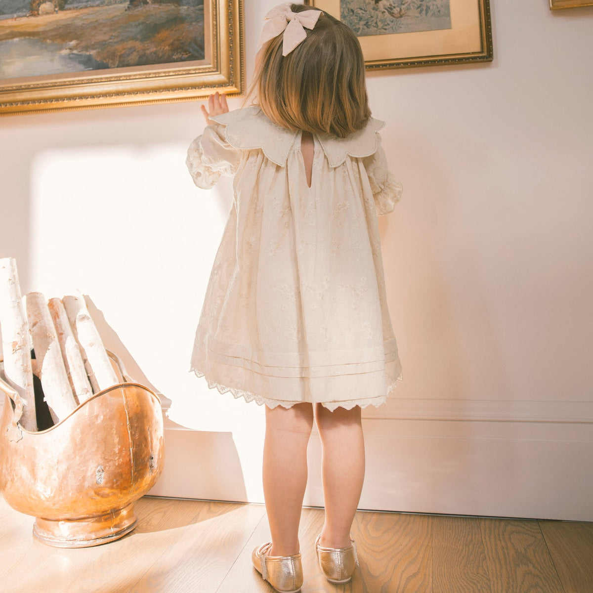 A young girl with a pink bow in her hair stands with her back to the camera, wearing a cream-colored lace dress and metallic gold shoes in a sunlit room with framed paintings and a copper pot.