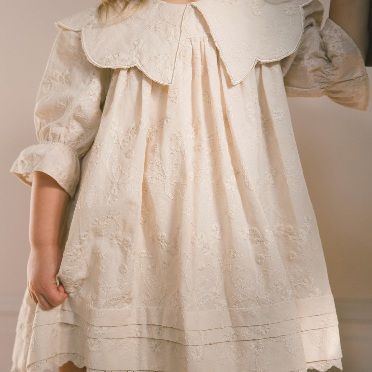 A cream-colored baby or toddler dress with a large ruffled Peter Pan collar, long puff sleeves, and delicate floral embroidery, shown on a child against a neutral background.