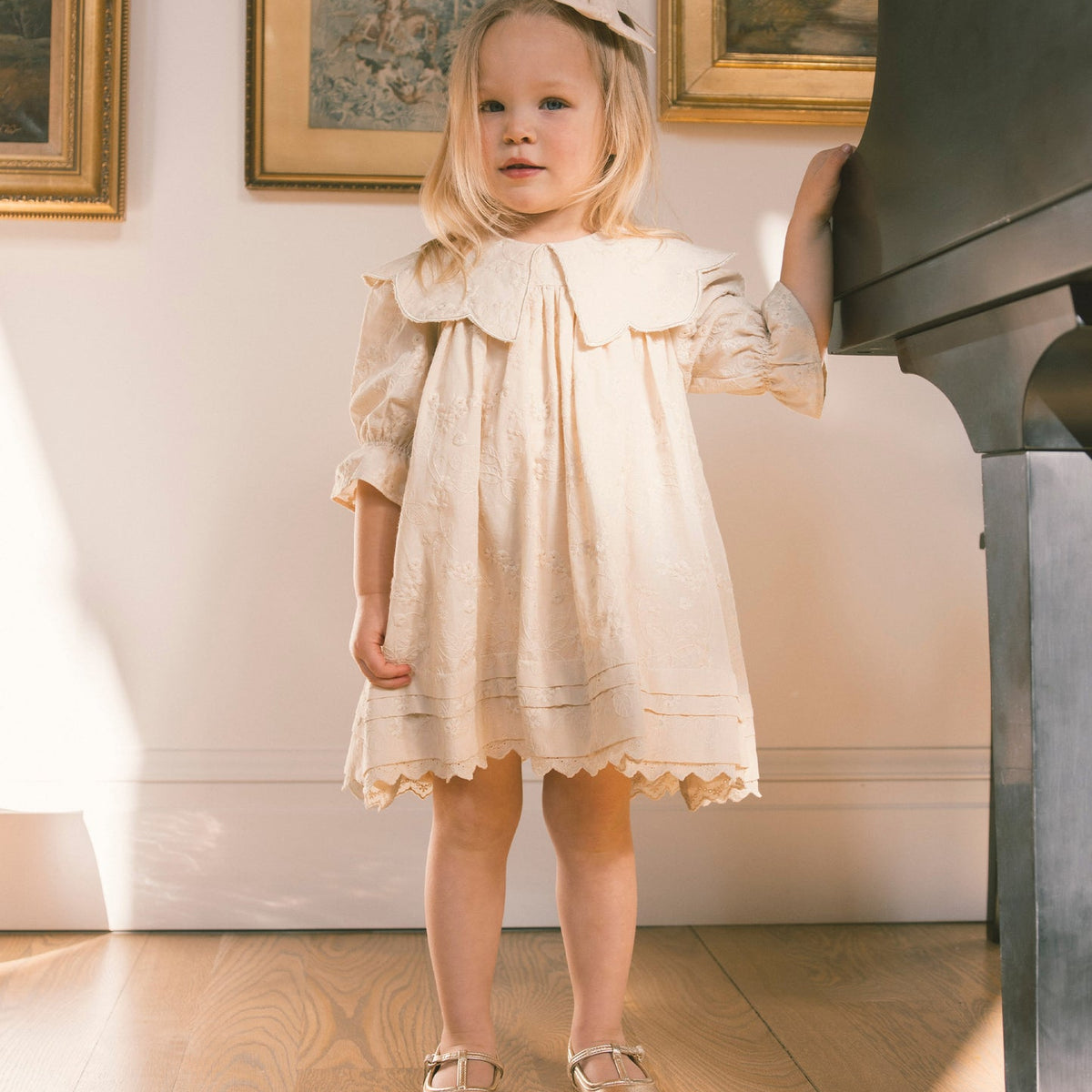 A young girl with blonde hair wearing a cream-colored lace dress and metallic gold shoes stands in a room next to a black grand piano, looking off-camera.