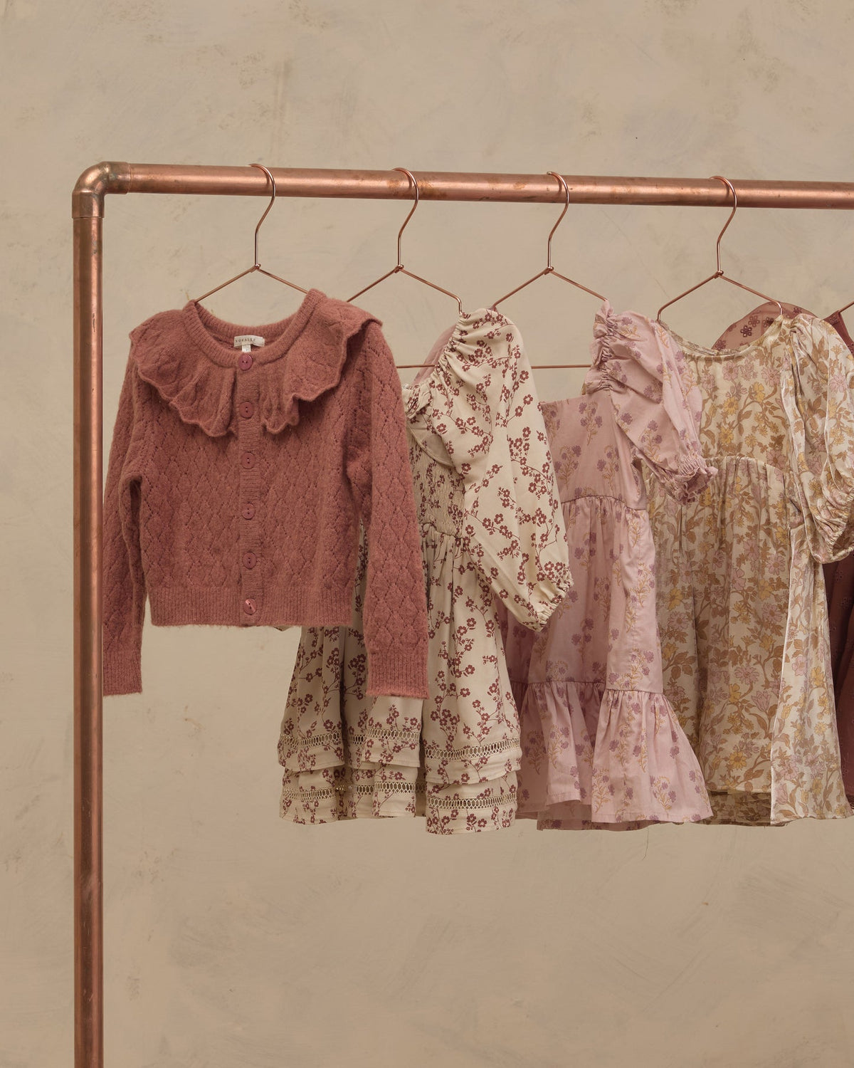 A copper-colored clothing rack displaying a collection of children's clothing, including a dusty rose knit cardigan with a ruffled collar and three floral dresses in cream, pink, and yellow tones, set against a textured beige wall.