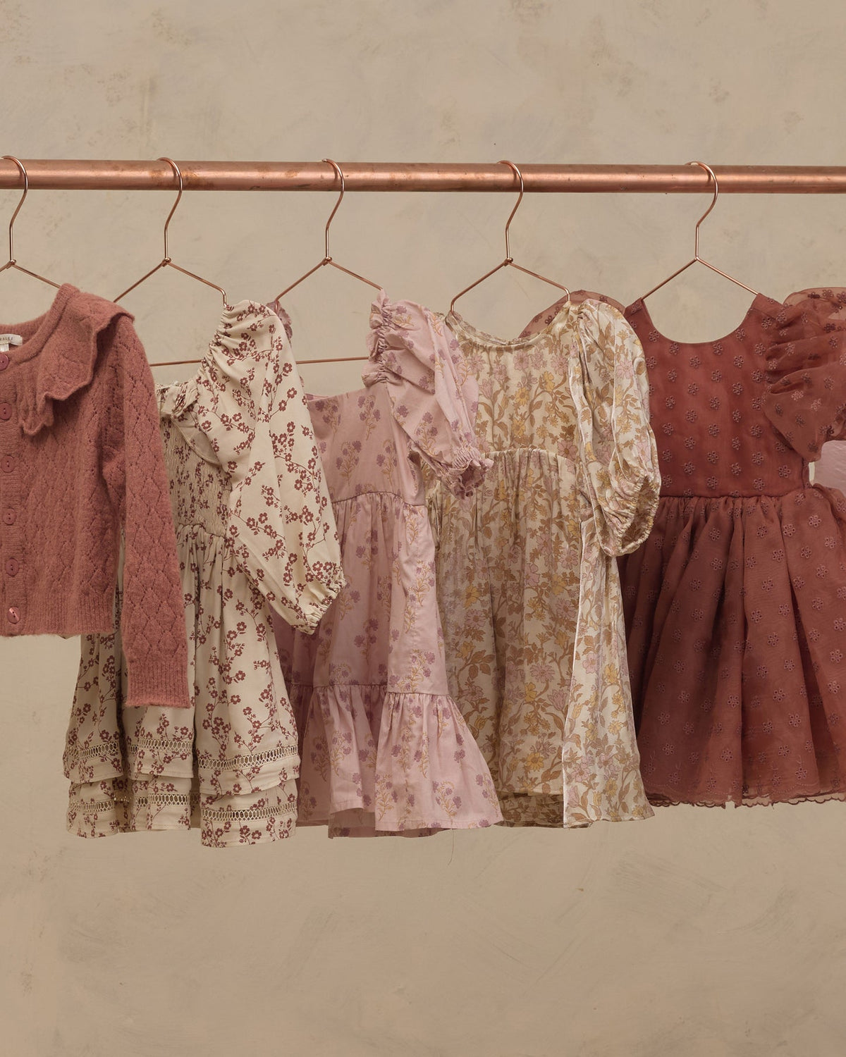 Four children's dresses in soft dusty pink, beige floral print, and rust tones hanging on a copper-colored clothing rack against a textured beige wall.