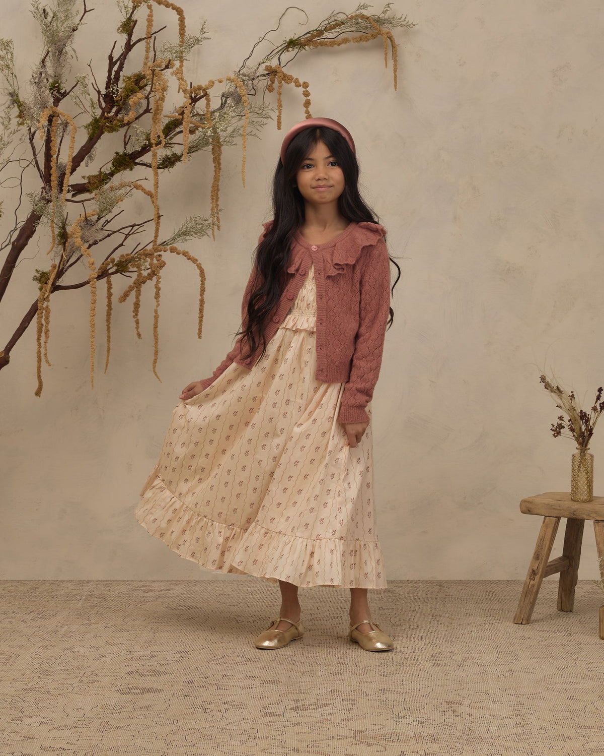 A young girl stands in a studio setting wearing a long cream dress with small pink floral prints, a dusty rose cardigan, and gold shoes. She is posed in front of a textured beige wall with dried botanical branches and a small wooden stool.