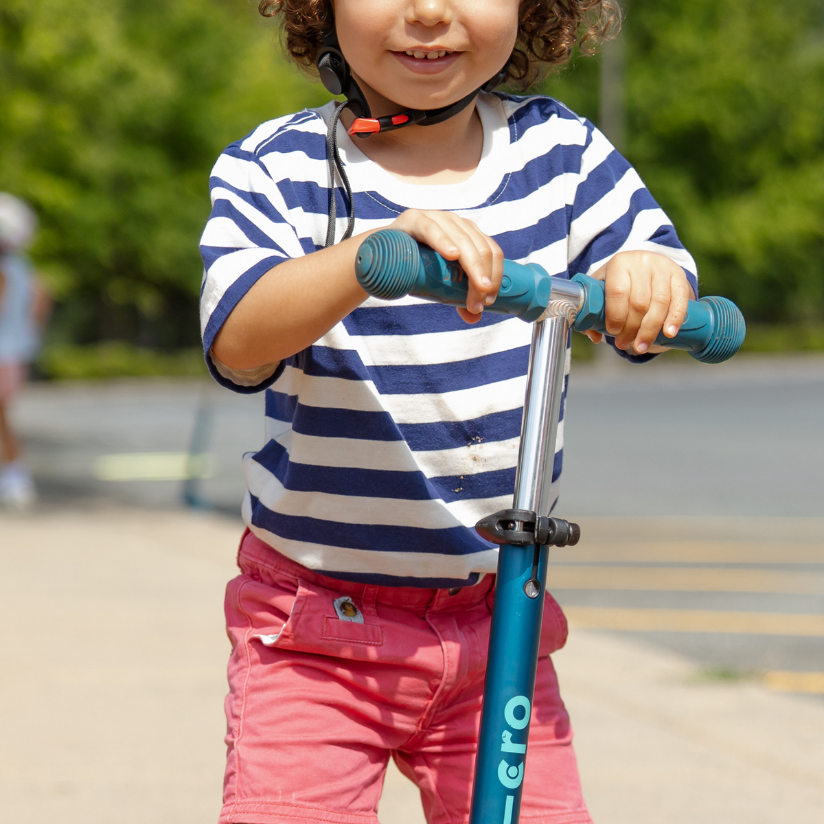 Child in a striped shirt and pink pants riding a blue scooter outdoors.