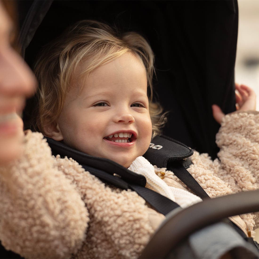 Child in a car seat wearing a beige coat, smiling.