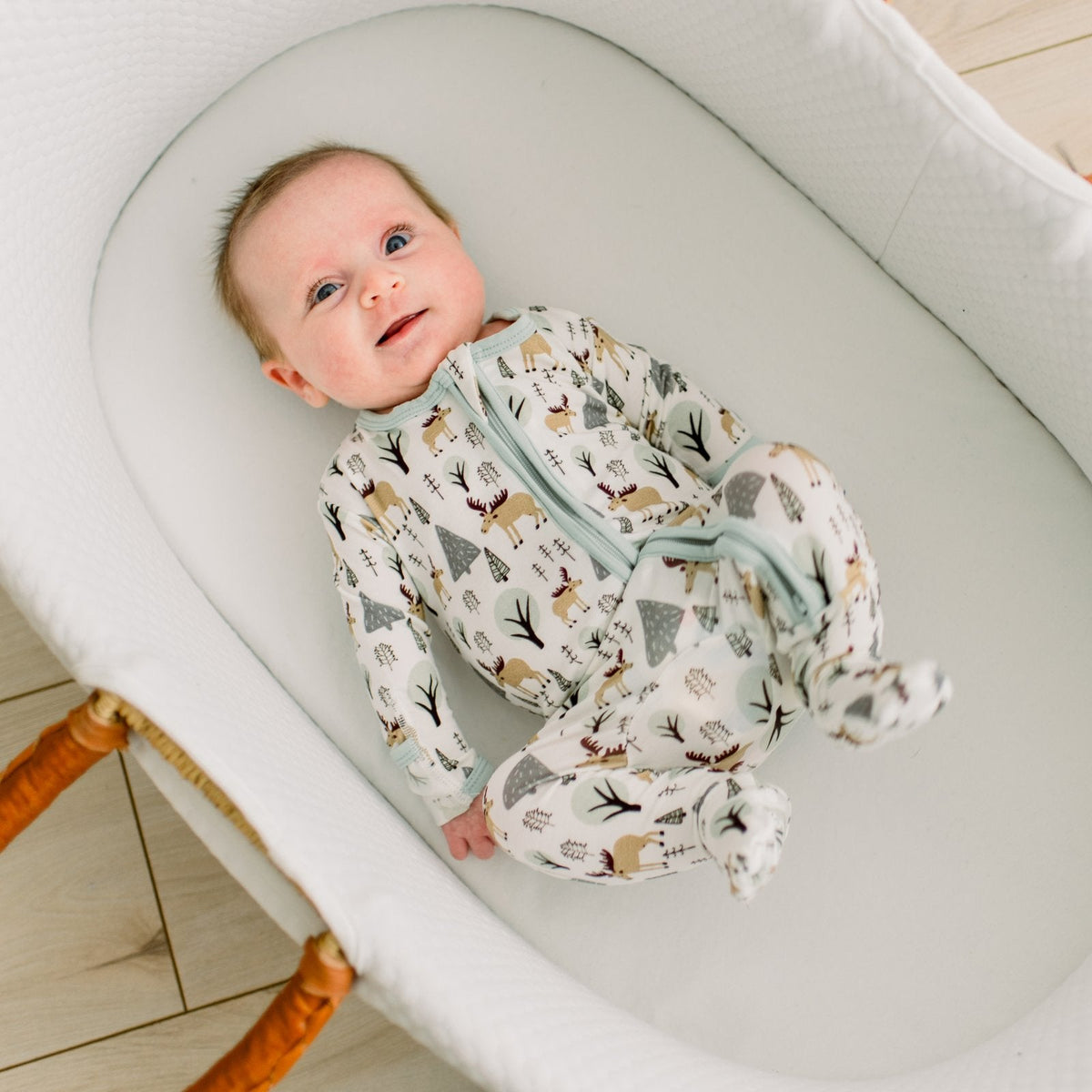 A baby lying on their back inside a white bassinet, wearing a footed one-piece sleeper with a woodland forest print in beige, brown, and gray with a light blue zipper trim.