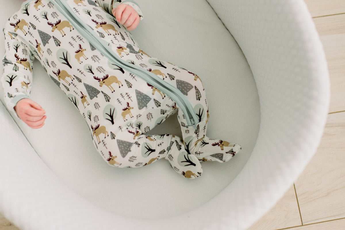 A baby wearing a long-sleeve zip-up sleep sack with a woodland animal print, lying inside a white bassinet.