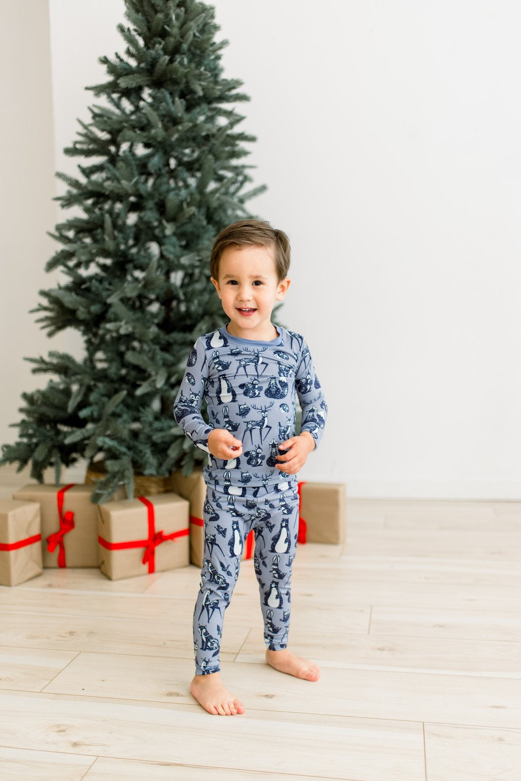 A young boy stands barefoot on a light wood floor in front of a Christmas tree and wrapped gifts, wearing a matching long-sleeve pajama set with a penguin print in blue and white.