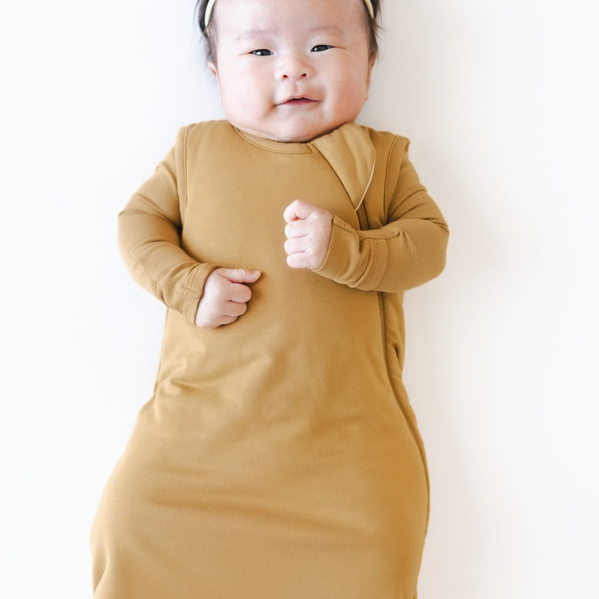A baby wearing a mustard yellow long-sleeve wrap swaddle and a matching bow headband, lying on a plain white background.