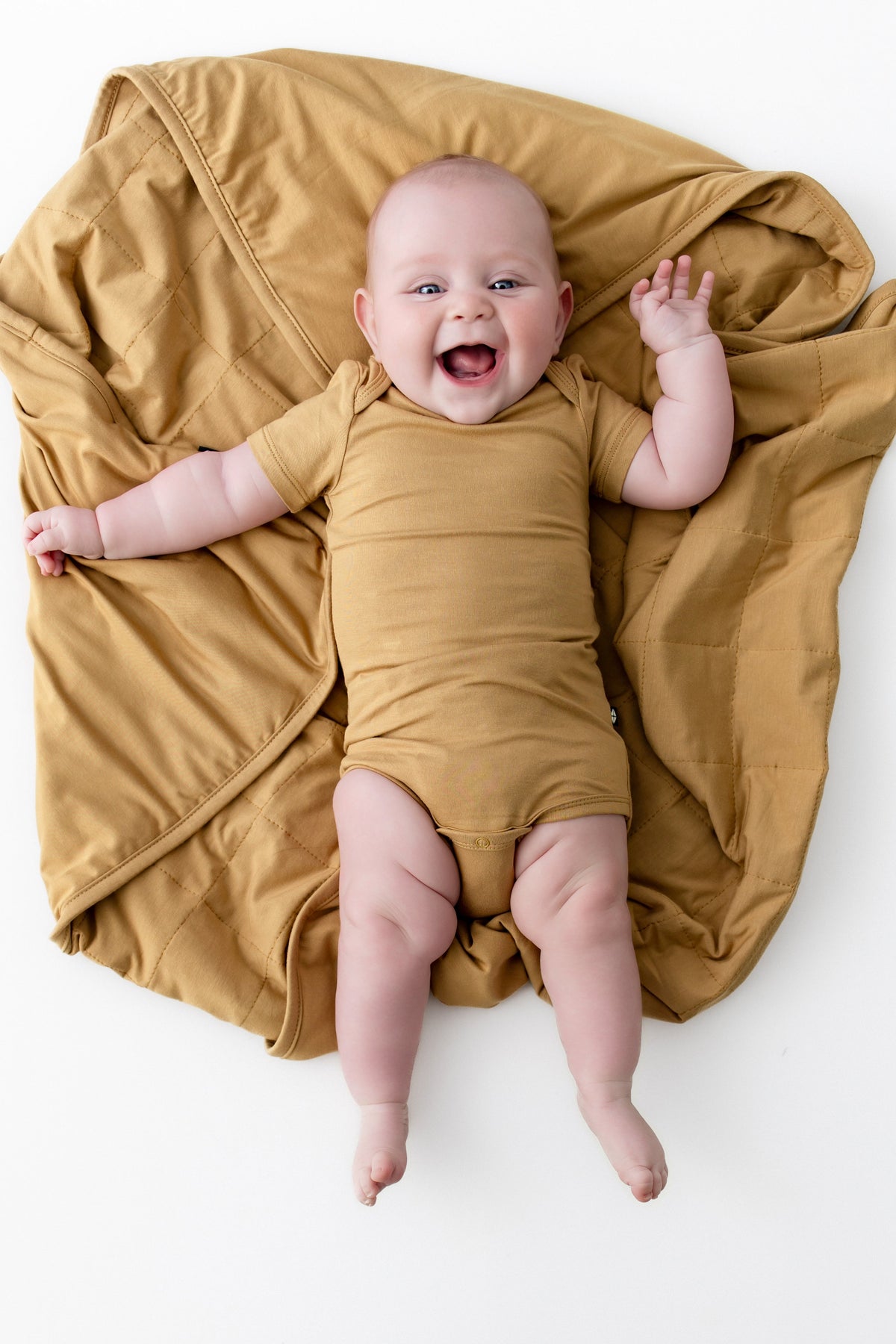 A smiling baby lying on their back on a white background, wearing a matching mustard yellow short-sleeve bodysuit and wrapped in a larger mustard yellow blanket.