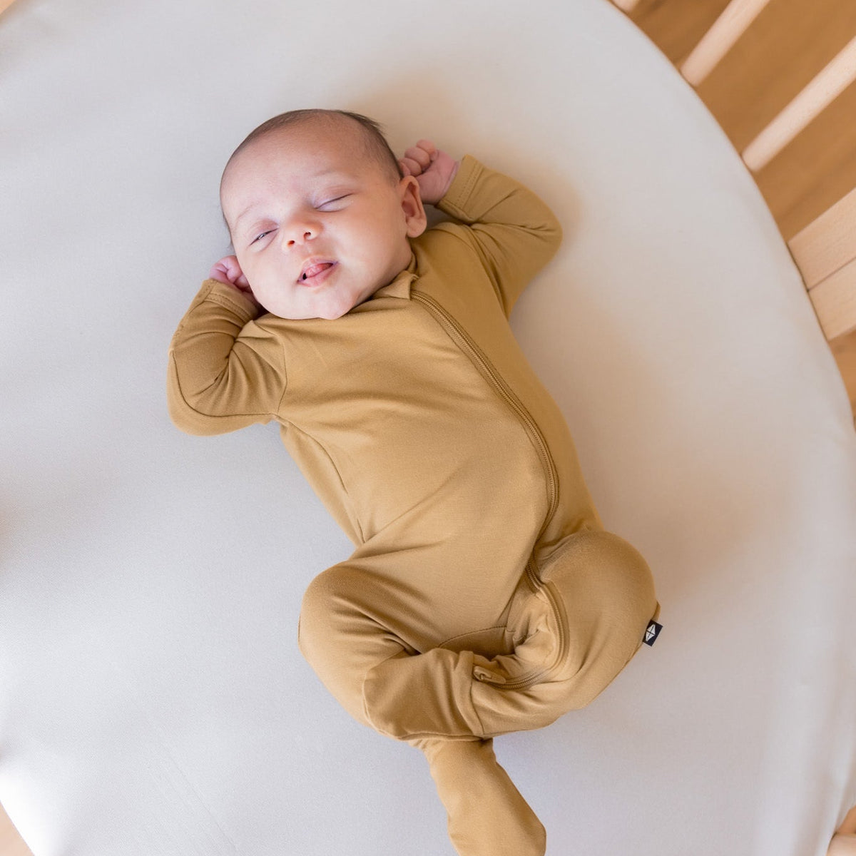 A sleeping newborn baby wearing a mustard yellow footed zip-up sleeper, lying on a white oval mattress inside a wooden crib.