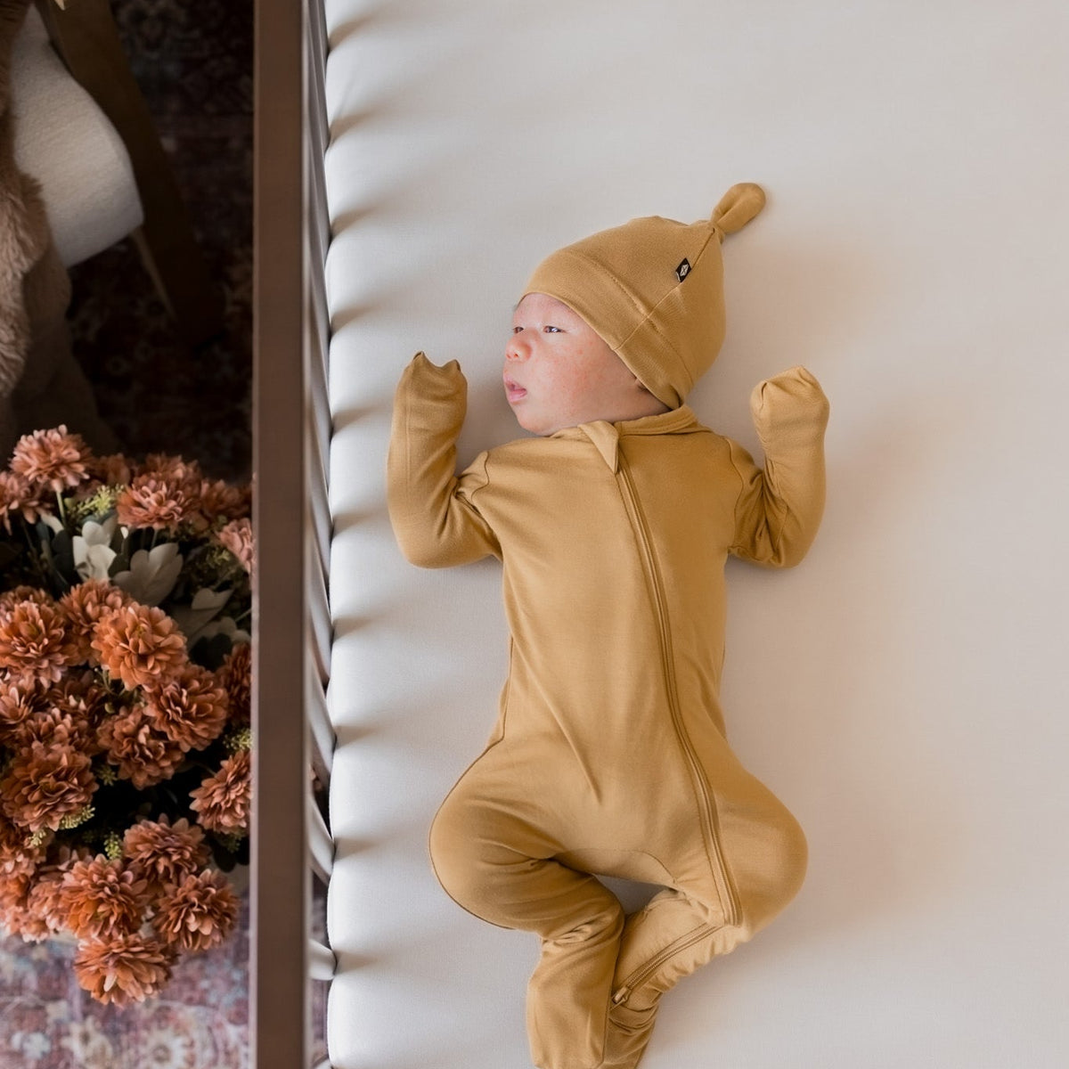 A newborn baby wearing a mustard yellow full-body footed onesie and matching hat, lying on their back on a white textured blanket next to a wooden frame with dried flowers.