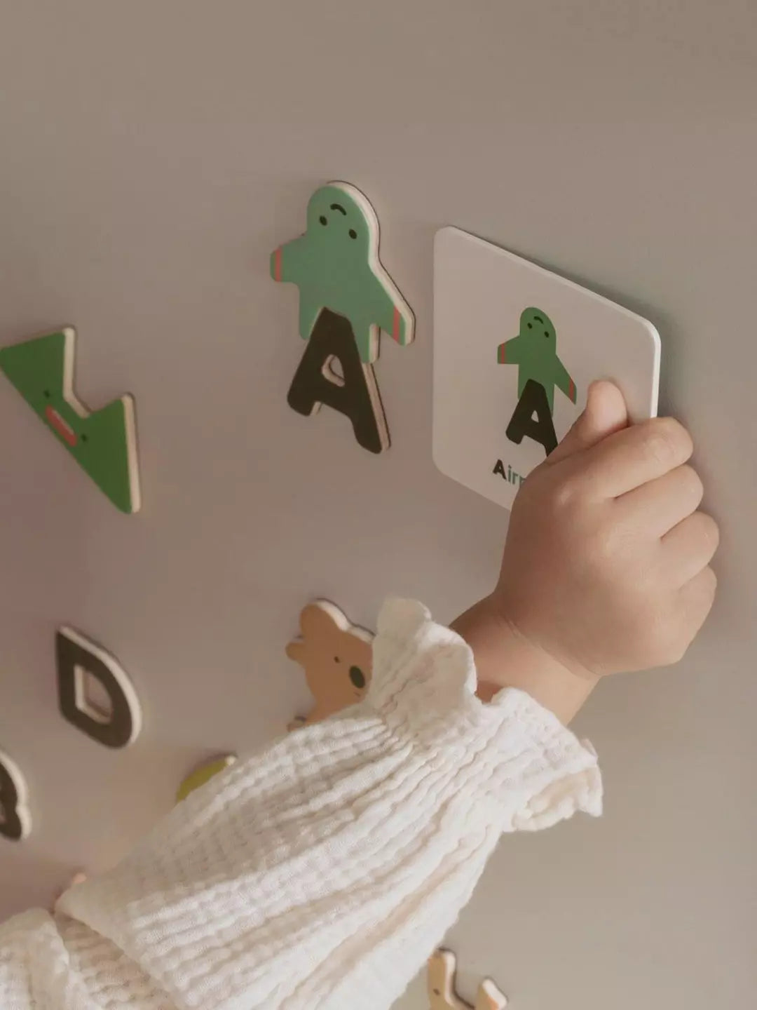 Child's hand holding a letter card with a green character on a wall with other letter cards.