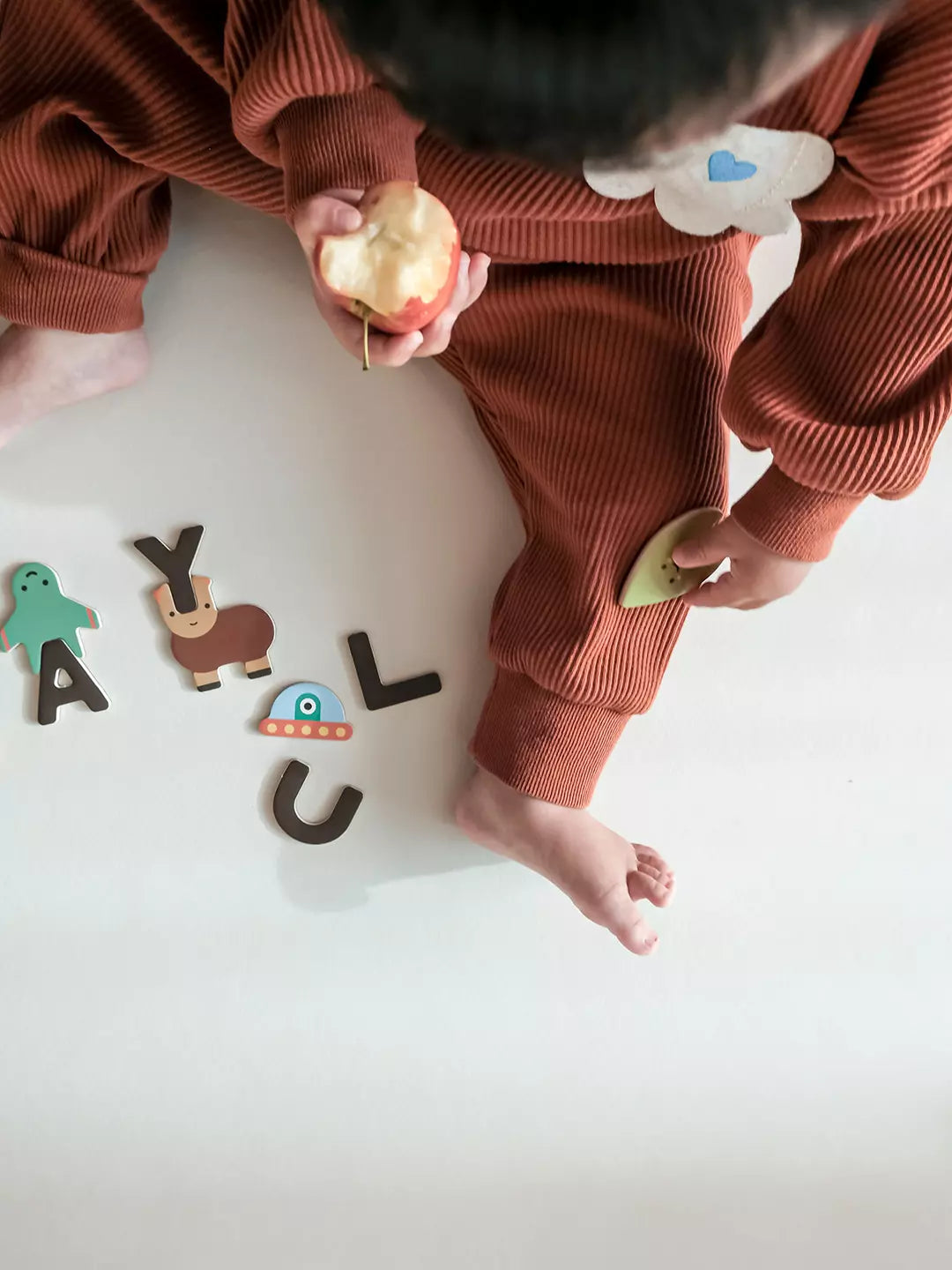 Child in brown outfit holding an apple with colorful letters on a white floor