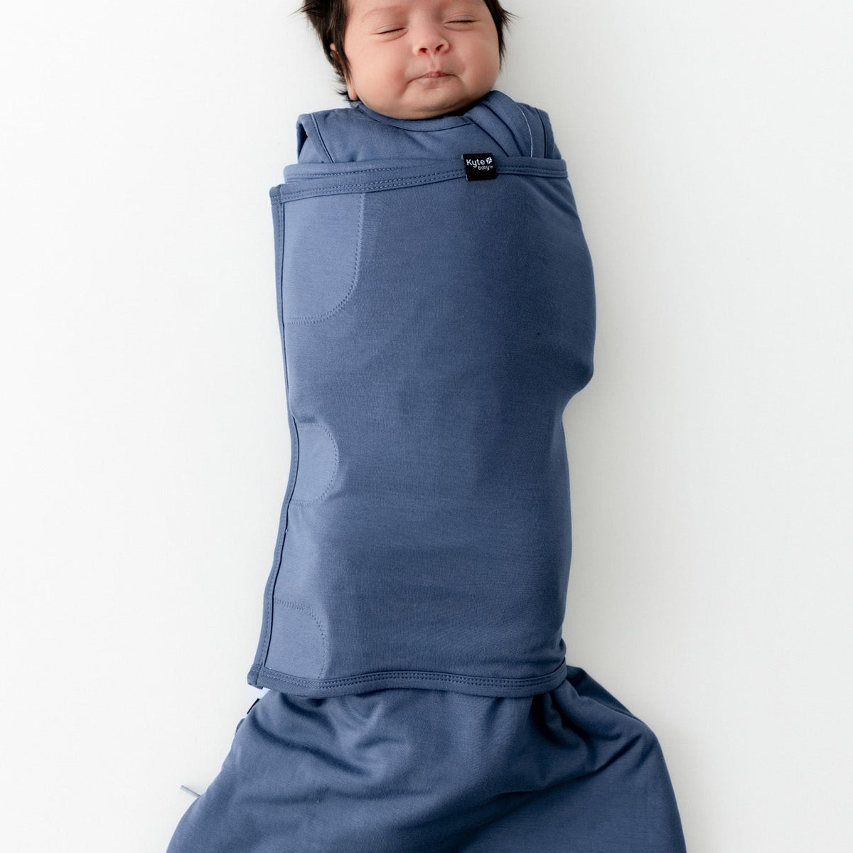 A sleeping newborn baby swaddled in a dusty blue wrap blanket, lying flat on a clean white background. The blanket is secured with a small black brand tag that reads 'MAMmoth'.