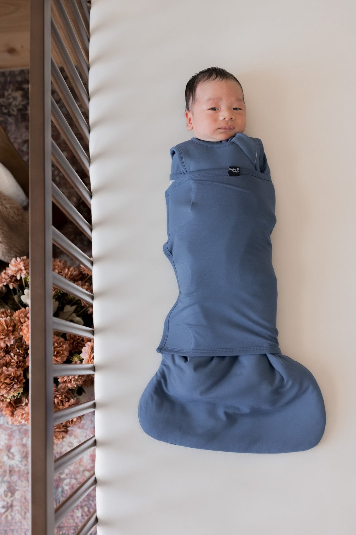 A newborn baby is lying on their back inside a crib, swaddled in a dusty slate blue wrap blanket. The brand tag on the chest of the wrap reads 'Mamaweb'.