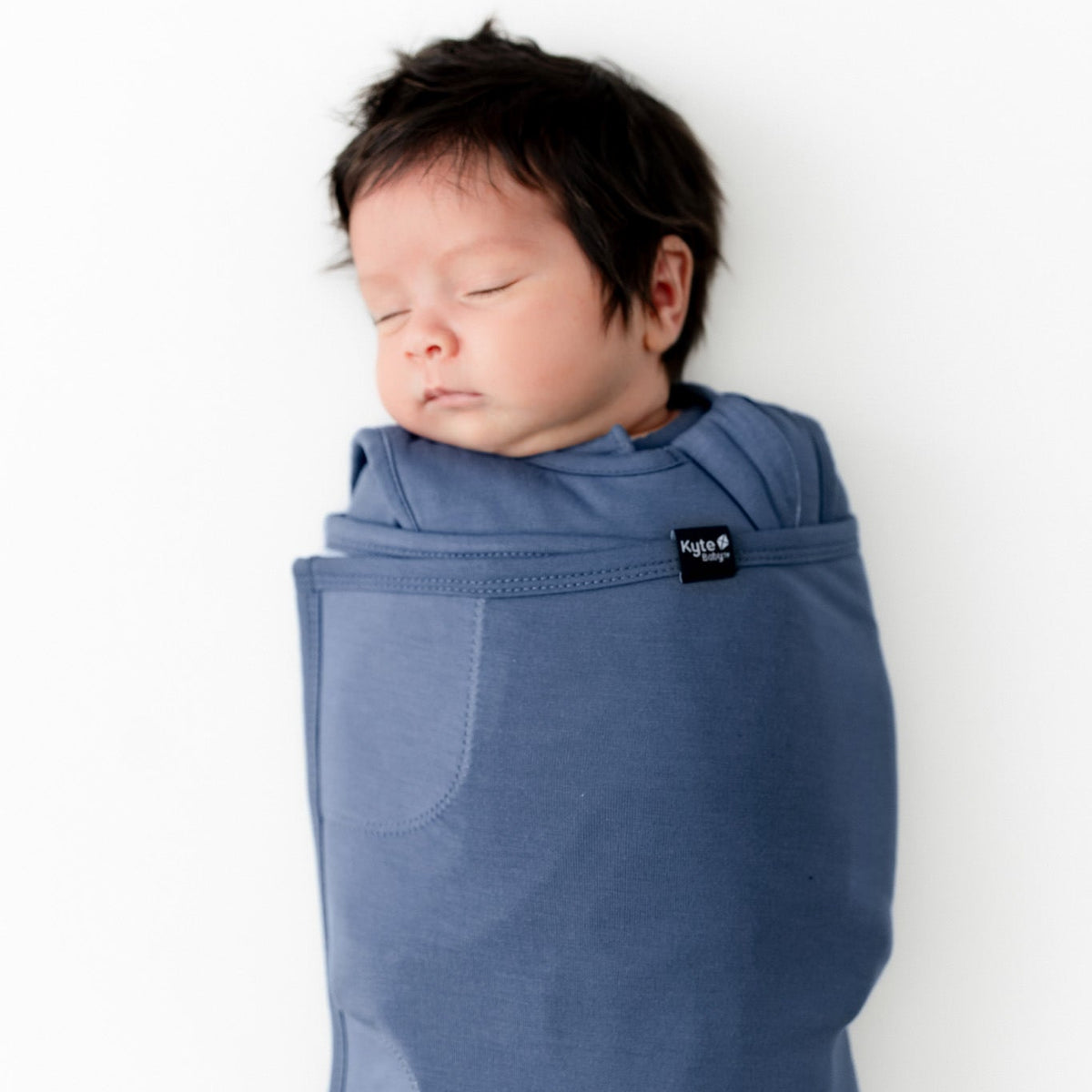 A sleeping newborn baby wrapped in a dusty blue fabric baby carrier wrap, shown against a plain white background.
