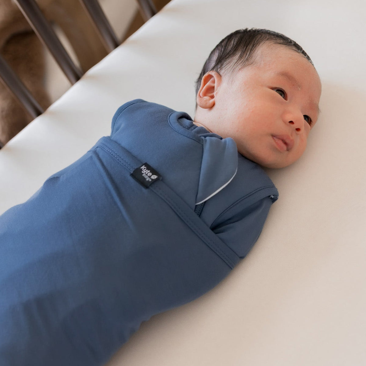 A newborn baby lying on a white mattress, swaddled in a blue wrap blanket. The baby is wearing a matching blue one-piece outfit with a small black brand tag that reads 'baba&boo'.