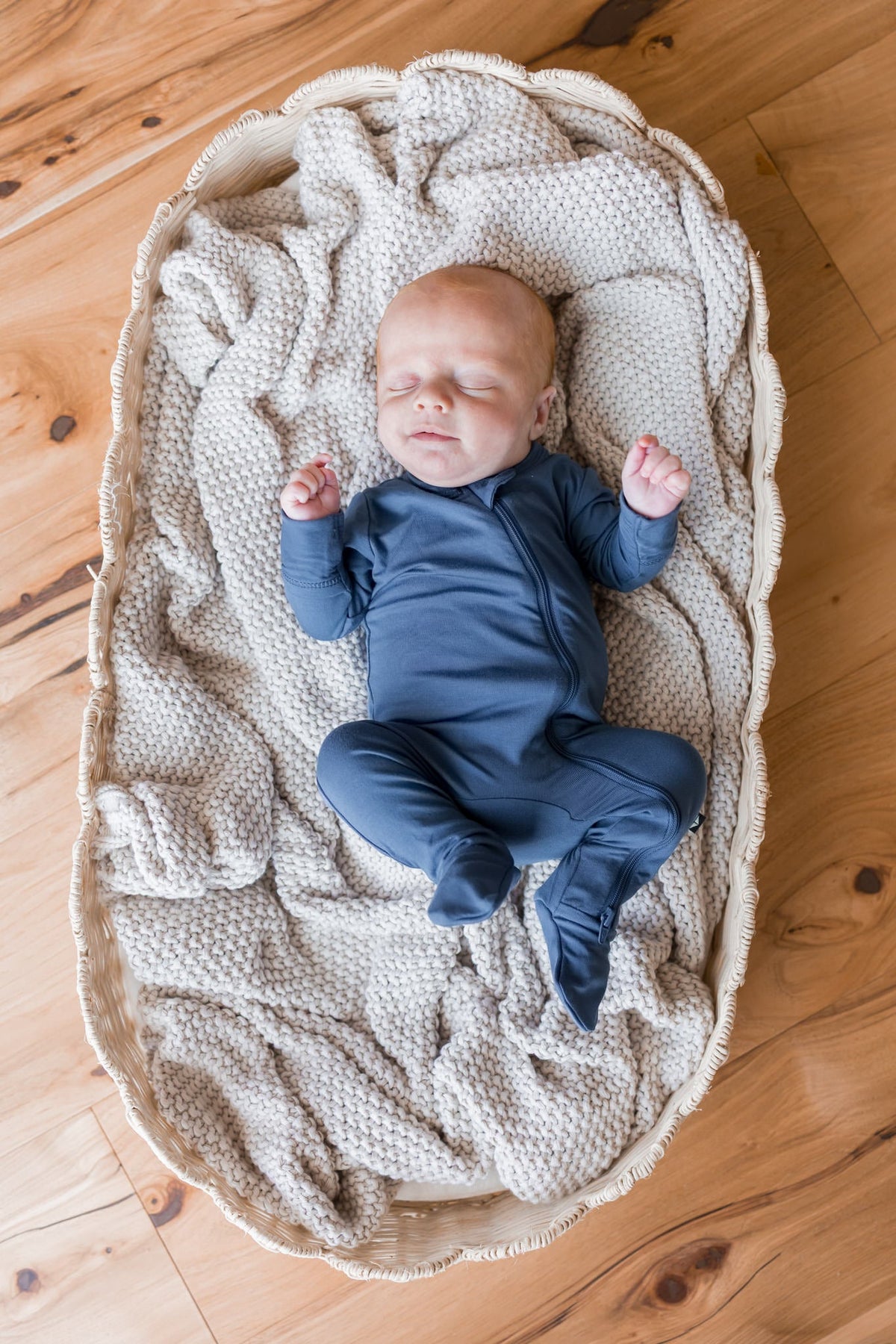 A sleeping newborn baby wearing a navy blue footed one-piece outfit, lying on a textured cream knit blanket inside a woven oval bassinet on a wooden floor.