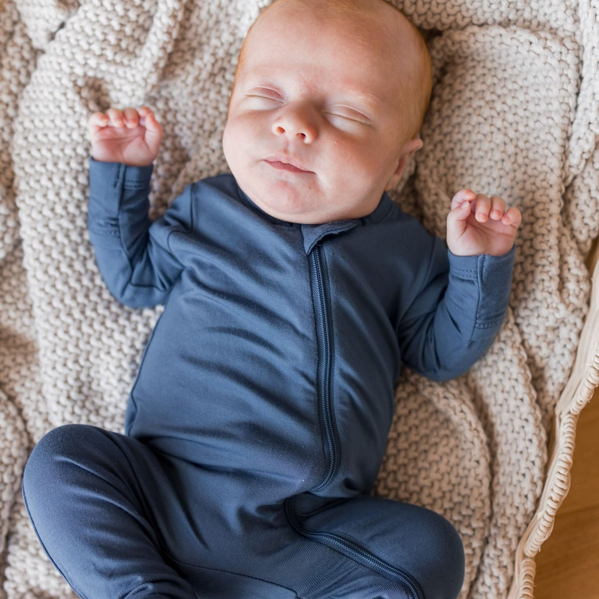 A sleeping newborn baby wearing a navy blue footed zip-up onesie, lying on a beige knit blanket inside a wicker basket.