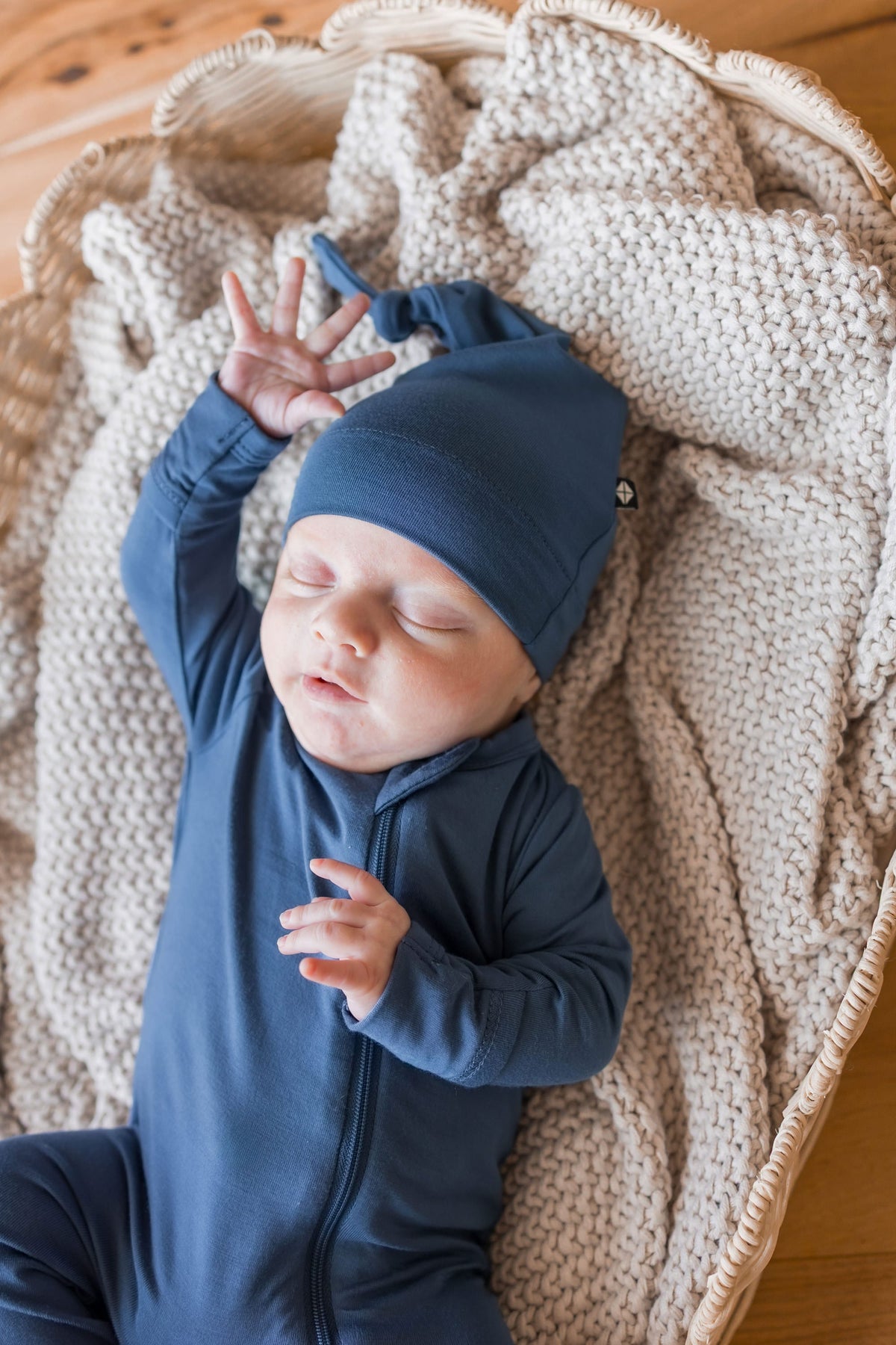 A sleeping newborn baby wearing a navy blue long-sleeve footless onesie and matching knit hat, lying on a beige chunky-knit blanket inside a wicker basket on a wooden floor.