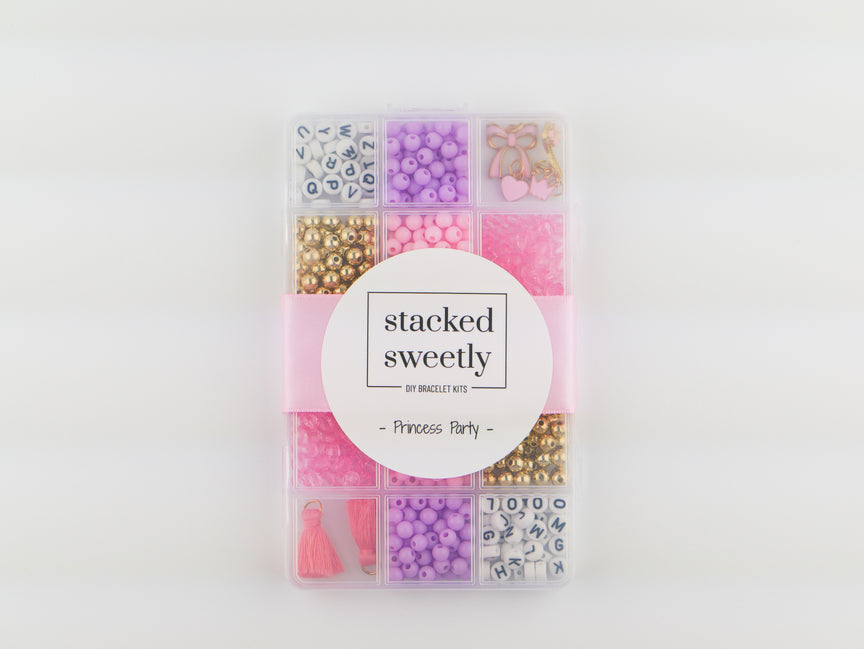 Princess Party DIY Bracelet Kit storage box shown from above, featuring sorted compartments of pink, purple, and white beads, alphabet letter beads, and gold charms with a central "stacked sweetly" band.